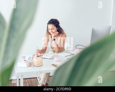 Cute young lady smiling and making notes in notepad while sitting at table in stylish office Stock Photo