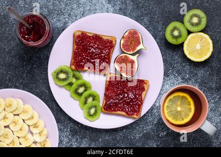 Toast sandwiches with raspberry jam, top view. Red confiture spread on a slice of white bread. Tea with lemon and fresh fruits on a dark board. Breakf Stock Photo