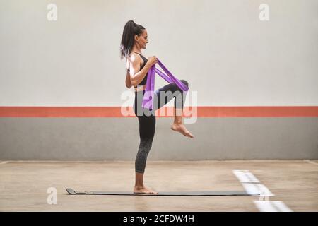 Cheerful athletic female in activewear standing barefoot on mat and stretching legs with resistance band during training