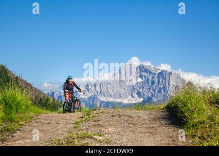 woman pedaling on a gravel road, in the background  the Civetta mountain near Cherz alm, tour with e-bike in the natural landscape of Dolomites, Stock Photo