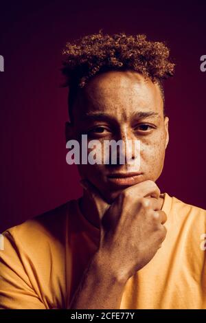 Freestyle. Mulatto freckled guy standing isolated on dark red in yellow light touching chin thoughtful close-up Stock Photo
