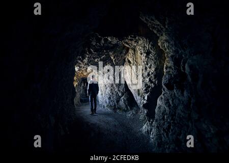 Young man is looking into light at the end of the tunnel. Young hiker walking  through tunnel in rock. Stock Photo