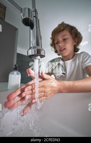 From below boy with towel on shoulder washing hands under running water carefully in modern bathroom at home Stock Photo