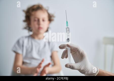 Crop hand in latex glove of anonymous doctor demonstrating syringe with vaccine medication before giving injection to boy Stock Photo