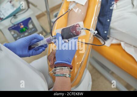 From above of crop female nurse in protective gloves with bag of blood in hand working with patient during procedure of blood donation in modern medical center Stock Photo