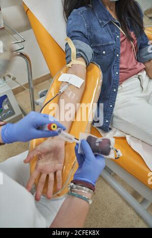 From above of crop female nurse in protective gloves with bag of blood in hand working with patient during procedure of blood donation in modern medical center Stock Photo