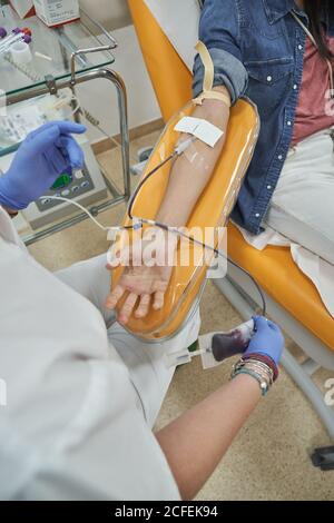 From above of crop female nurse in protective gloves with bag of blood in hand working with patient during procedure of blood donation in modern medical center Stock Photo