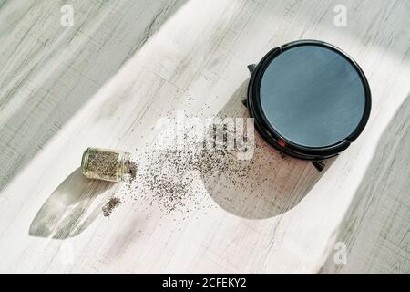 From above of round robotic vacuum cleaner sliding on light laminate floor and removing  dirt spilled out of glass pot Stock Photo