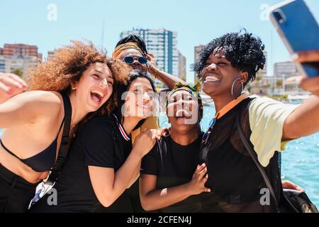 Group of charismatic multiracial girlfriends standing at waterfront and taking selfie on smartphone during stroll in summer Stock Photo
