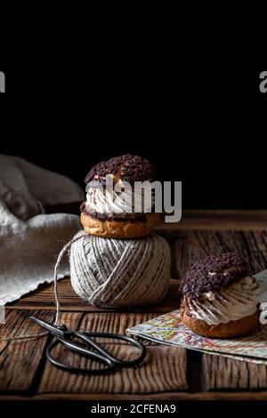 Homemade choux pastries with cream and chocolate scissors and ball of yarn arranged on texture wooden surface against black background Stock Photo