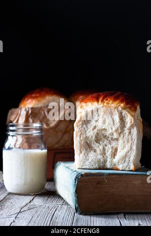 Small jar of fresh milk and buns of fresh bread placed on timber table near old recipe book against black background Stock Photo