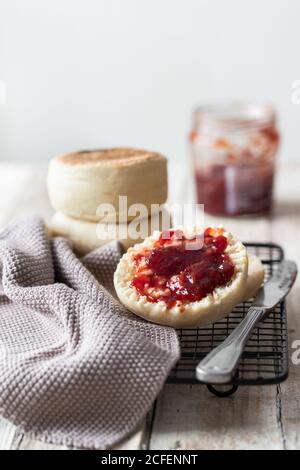 Appetizing freshly baked bun cut in half with homemade strawberry jam placed on metal grid with gray cloth and knife on kitchen table Stock Photo