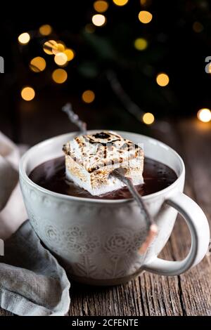 White cup with tasty hot chocolate drink garnished with roasted marshmallow placed on wooden table against dark background with sparkles