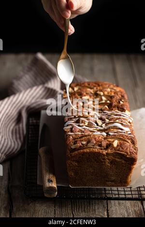 Crop housewife with spoon in hand pouring white sugar icing over homemade banana bread with nuts placed on metal grid on wooden table Stock Photo