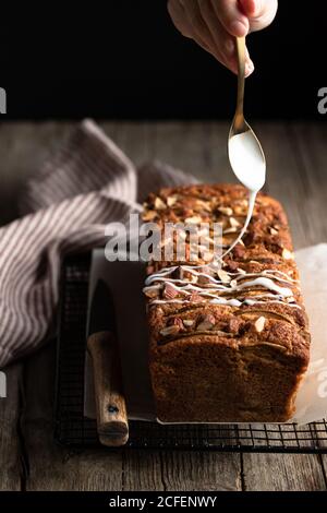 Crop housewife with spoon in hand pouring white sugar icing over homemade banana bread with nuts placed on metal grid on wooden table Stock Photo