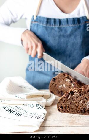 Hands of Woman in apron holding long steel knife and cutting rye bread with raisins and nuts Stock Photo