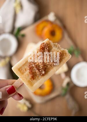 Hand of anonymous female holding bitten coconut and pineapple ice cream over blurred table Stock Photo