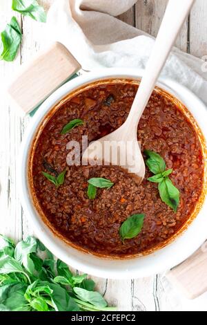 From above white ceramic pan with ragout Bolognese and spoon placed on wooden table with towel and basil Stock Photo