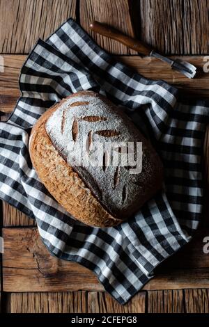 Top view of loaf of fresh spelt bread placed on checkered napkin near tool on lumber table Stock Photo