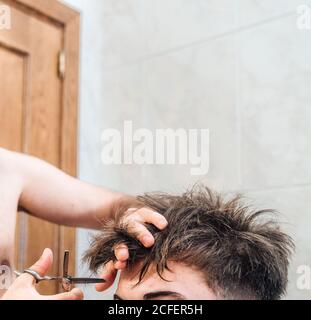 unrecognizable male making haircut to guy using scissors against blurred interior of light bathroom at home Stock Photo