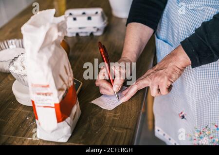 From above of crop anonymous mature female in apron making calculation on paper while preparing ingredients for pastry in home kitchen