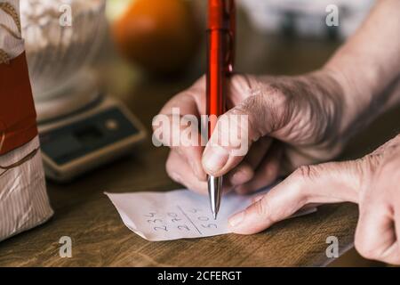 From above of crop anonymous mature female in apron making calculation on paper while preparing ingredients for pastry in home kitchen Stock Photo