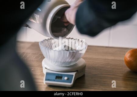 Cropped unrecognizable female hand holding sugar jar spilling and weighting sugar with electronic kitchen scale with glass bowl on top on wooden table Stock Photo