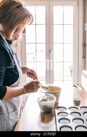 Side view of elderly housewife standing at table with muffin tray and pouring batter with spoon in cupcake liners Stock Photo
