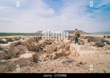 Side view of young man in yellow jacket and backpack with bicycle standing on stony hill stretchering high between stony hills in semi-desert Bardenas Reales Navarra Spain Stock Photo