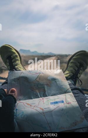 Road map in hands of man putting legs in green boots on dashboard of car moving along endless green fields in semi-desert Bardenas Reales Navarra Spain Stock Photo