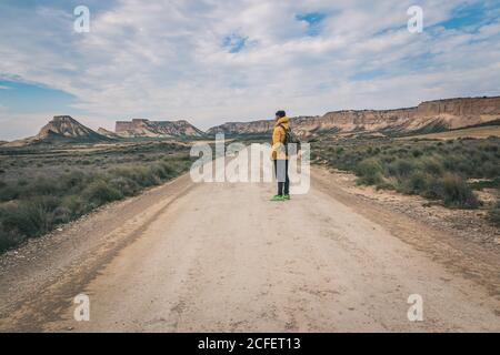 Side view of young man in yellow jacket and backpack standing on empty road stretchering high between stony hills in semi-desert Bardenas Reales Navarra Spain Stock Photo