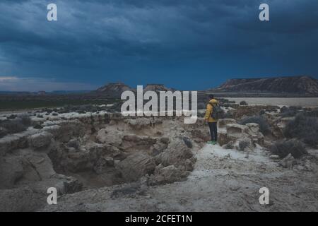 Side view of young man in yellow jacket and backpack standing on stone and looking at impressive blue sky in twilight in semi-desert Bardenas Reales Navarra Spain Stock Photo