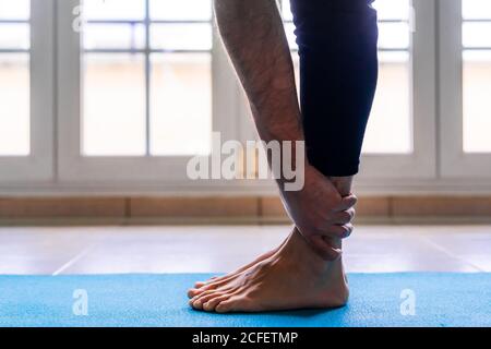 Low angle side view of unrecognizable flexible barefoot man in sportswear doing standing forward bend exercise while training alone on sports mat against window in light spacious room at home Stock Photo