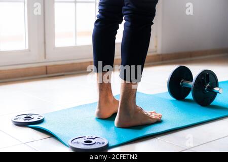 Low angle back view of crop barefooted male athlete in sportswear standing on bright blue sports mat against black collapsible dumbbell while preparing to workout in light spacious gym Stock Photo