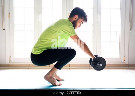 Side view of concentrated barefoot male athlete in sportswear doing squat exercise with collapsible dumbbell while training alone on sports mat against window in spacious workout room Stock Photo