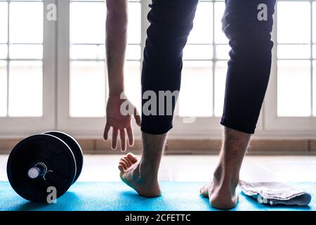 Back view of unrecognizable flexible barefoot man in sportswear doing standing forward bend exercise near dumbbell while training alone on sports mat against window in light spacious room at home Stock Photo