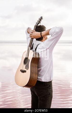 From behind man in white shirt holding acoustic guitar behind back while standing on shore on cloudy day in USA