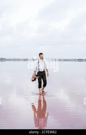 man in white shirt and suspenders holding guitar while standing barefoot in water by shore on cloudy day Stock Photo