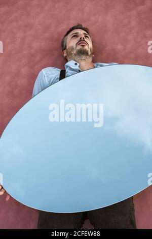 From above reflection of pensive man in shirt and suspenders lying down on pink water holding oval mirror reflecting blue sky Stock Photo