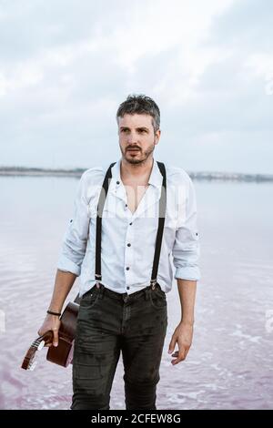 man in white shirt and suspenders holding guitar while standing barefoot in water by shore on cloudy day Stock Photo
