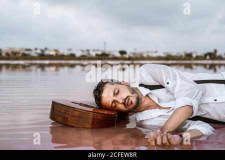 Sleepy man in white shirt and suspenders lying down with closed eyes with head resting at an acoustic guitar in sea at sandbank Stock Photo