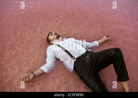 From above classy man in white shirt and suspenders lying on back and floating with outstretched hands in sea at sandbank Stock Photo