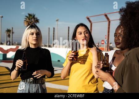 Serious youthful diverse female students resting and drinking alcohol while talking and standing on stadium in summertime Stock Photo