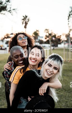 Multiethnic group of young laughing female friends in casual clothes having fun and cuddling with each one on lawn Stock Photo