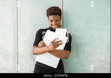 Cool African American smart Woman using a mobile phone to take a selfie while holding a laptop standing leaning on concrete wall Stock Photo