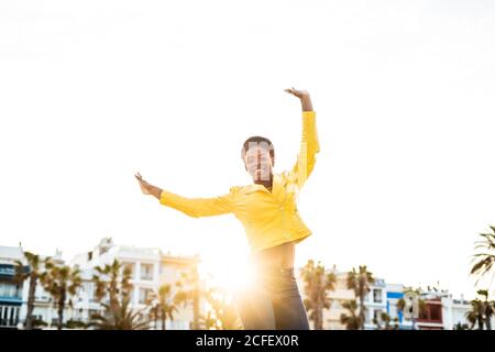 Low angle of happy African American Woman in stylish bright jacket jumping with hands up on white background Stock Photo