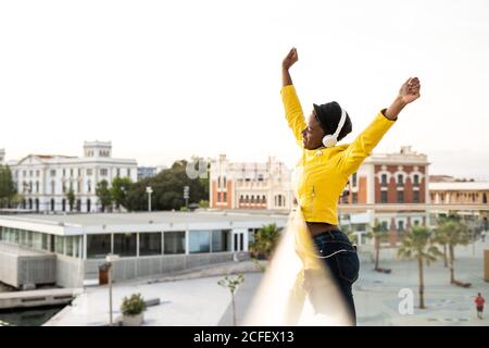 Side view content African American Woman in trendy jacket listening to music in headphones while leaning on glass balcony and looking away with arms up on blurred background Stock Photo