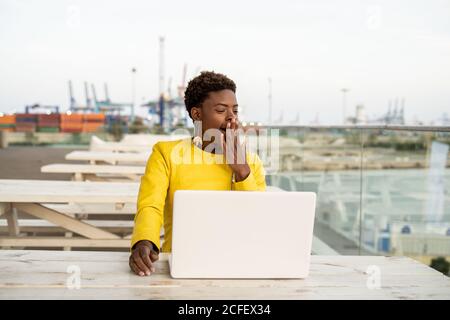 Tired African American Woman in yellow jacket yawning while using laptop at wooden desk in city on blurred background Stock Photo