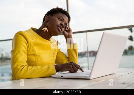 Tired African American Woman in yellow jacket yawning while using laptop at wooden desk in city on blurred background Stock Photo