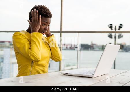 Surprised face of black African American Woman in yellow jacket using laptop at wooden desk in city on blurred background Stock Photo